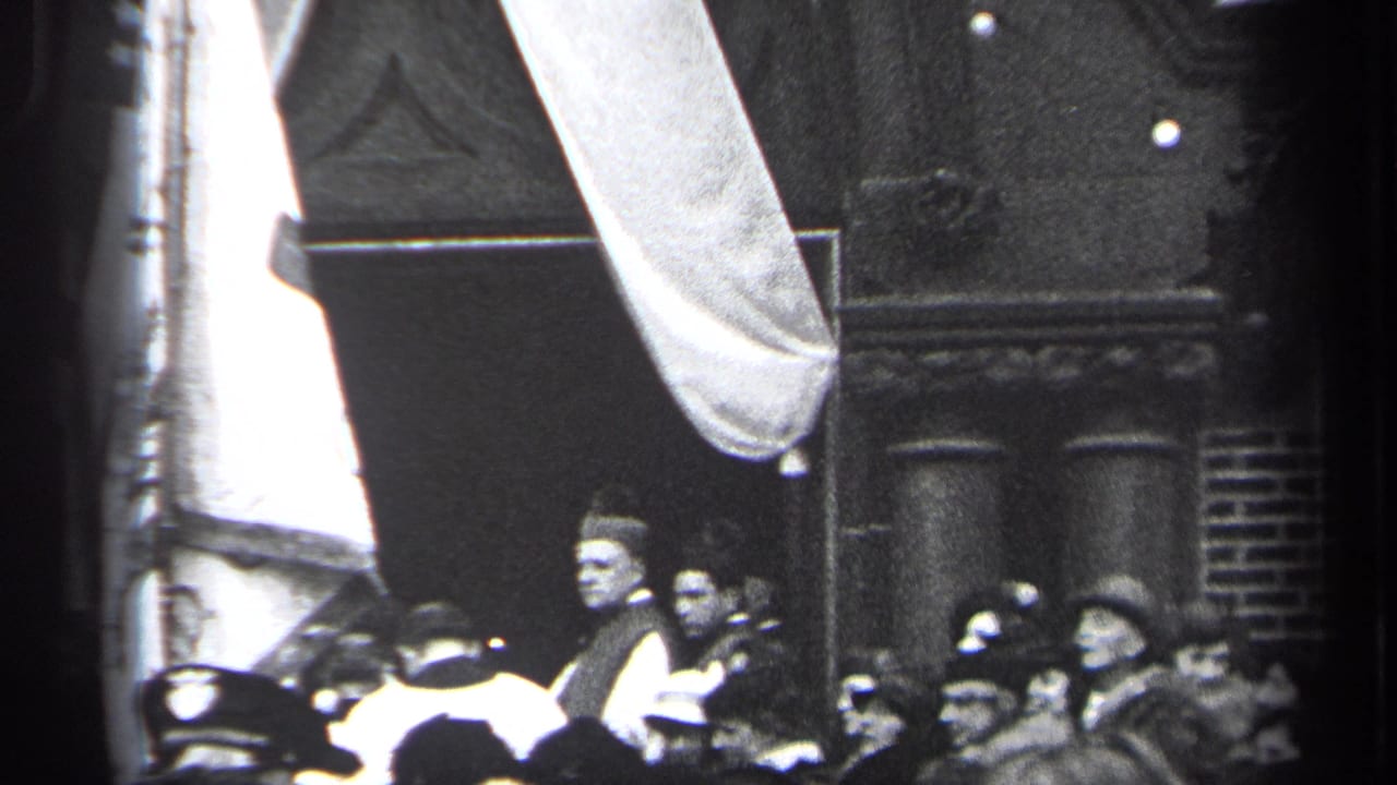 Crowd in hats watching a parade with a partial banner overhead.