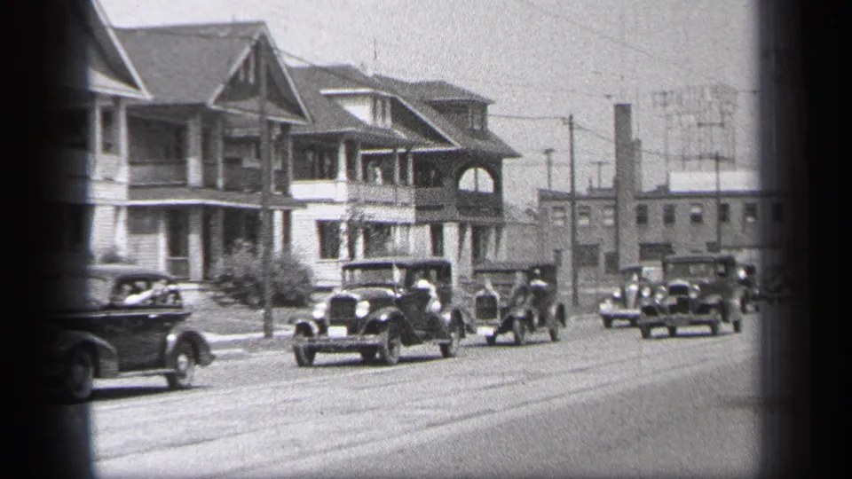 1937 Cleveland street view with 1930s cars moving in a line past porch-front houses and a small group of spectators.