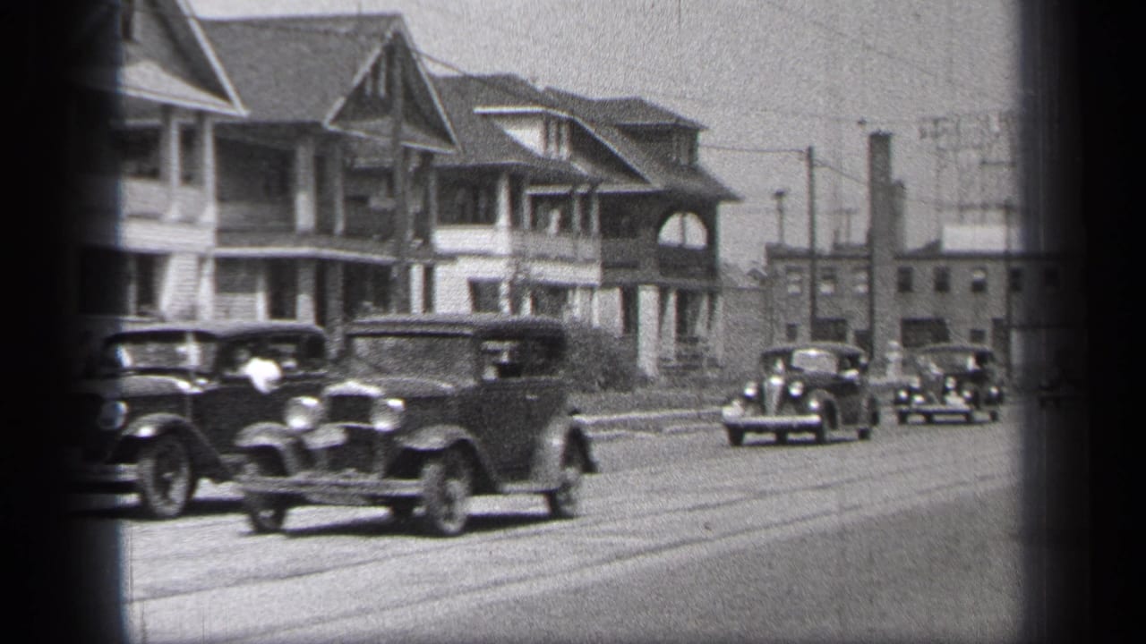 Vintage cars drive along a broad street past two-story houses and utility poles.