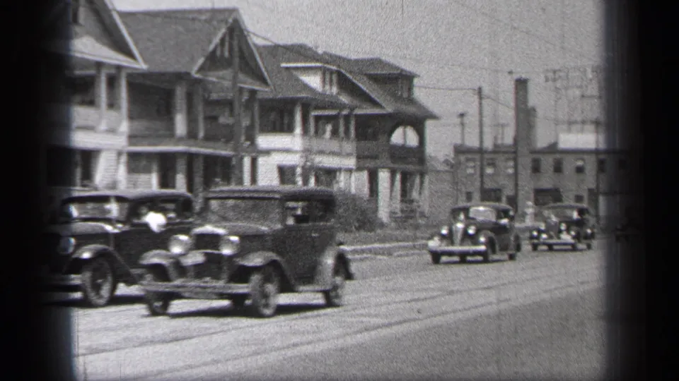 1937 Cleveland street view with several vintage automobiles passing two-story homes, overhead wires, and distant low buildings.