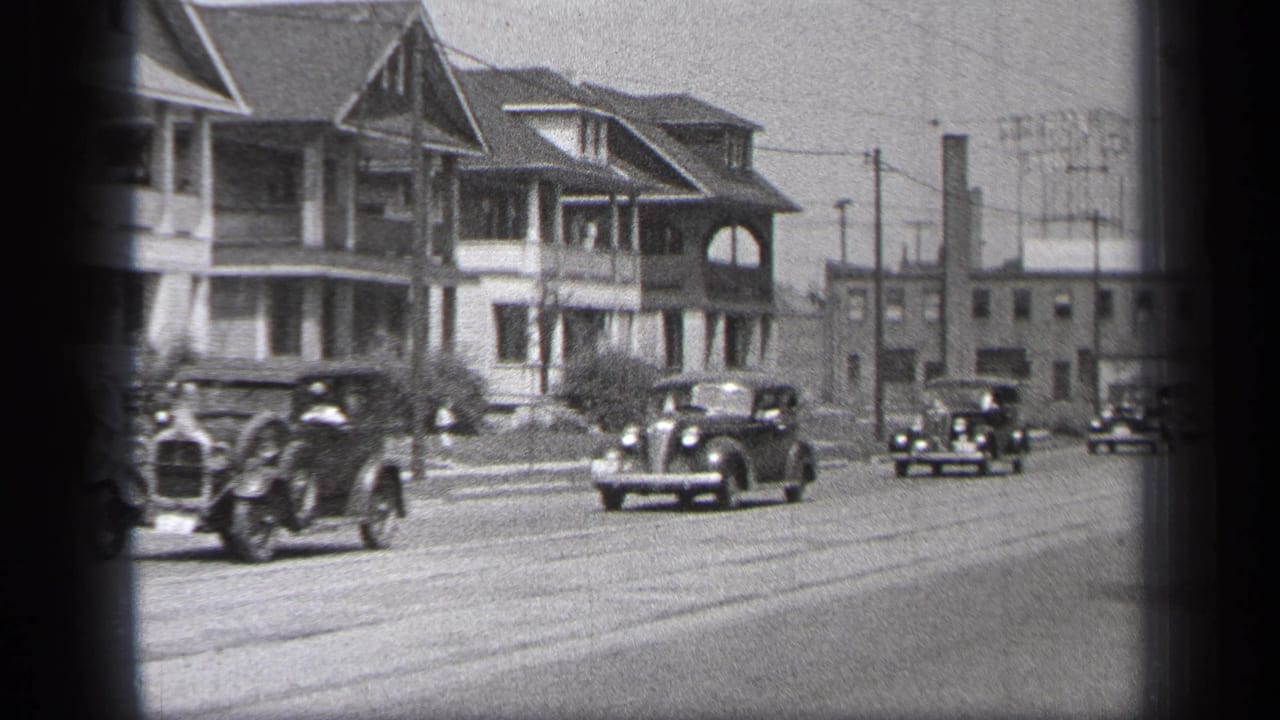 Several vintage automobiles approach and pass on a residential street with houses in the background.