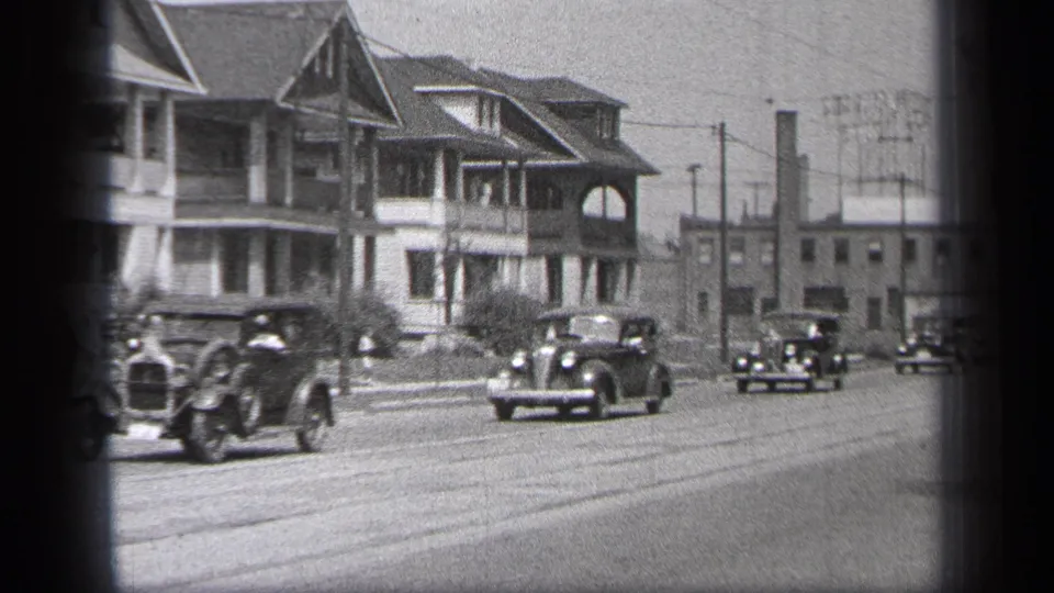 Vintage cars move through a Cleveland residential street scene, with porched houses, road markings, wires, and low buildings behind them.