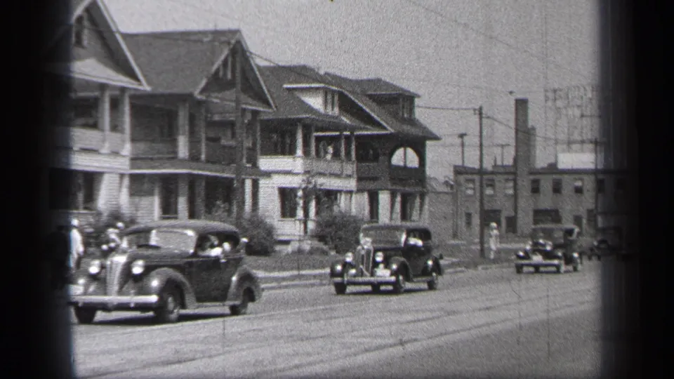 Late-1930s automobiles pass through a Cleveland residential block, framed by two-story homes, utility poles, and distant industrial-looking buildings.