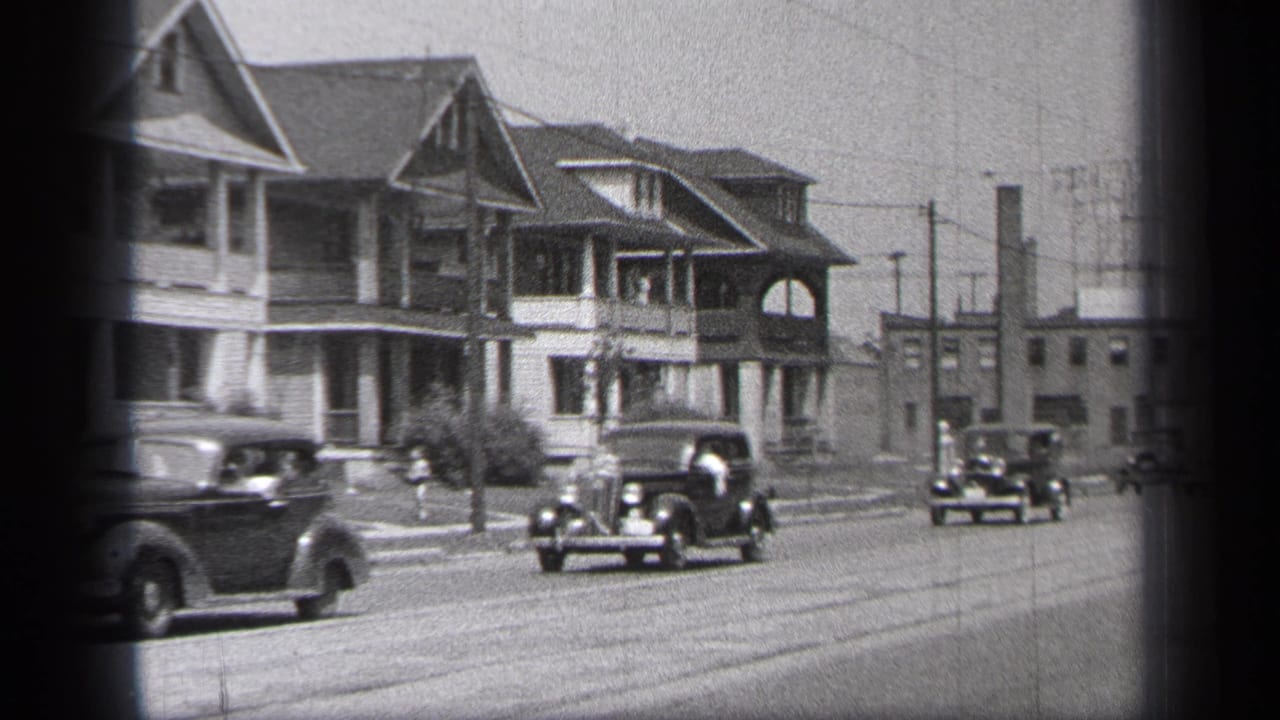 Classic cars pass porch-front houses on a wide residential street.