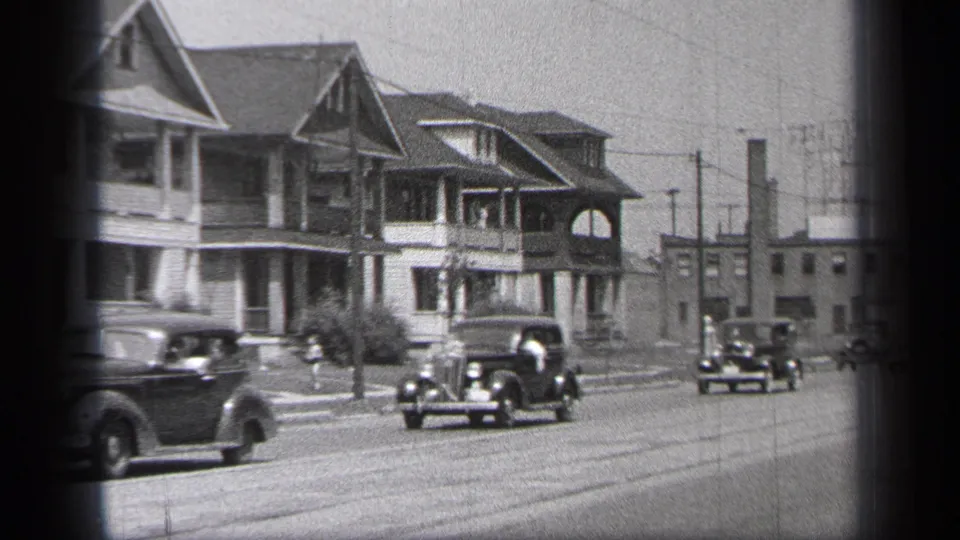 1937 Cleveland home-movie frame showing vintage cars moving past gabled houses, sidewalks, utility poles, and overhead wires.