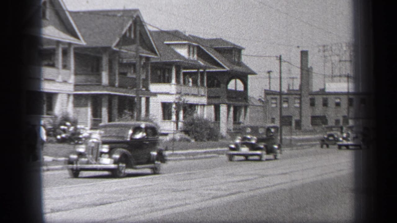 Several vintage cars drive past houses and utility poles.