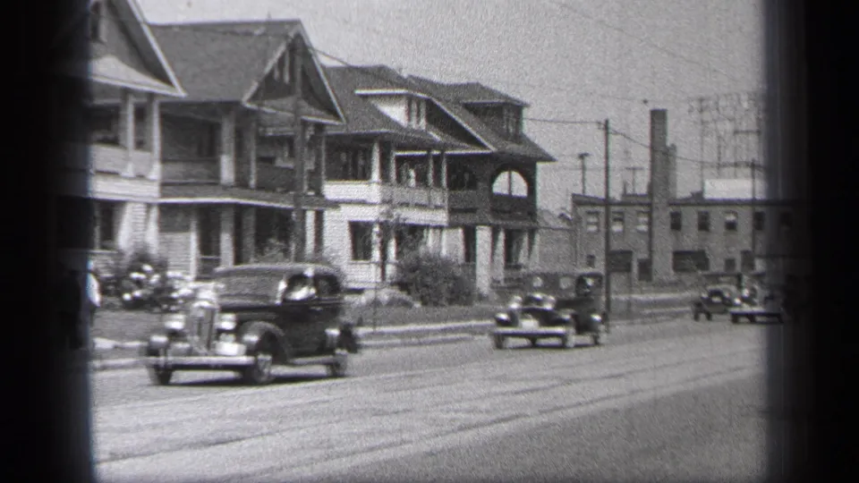 1937 Cleveland street view with multiple classic cars, porch-front homes, overhead lines, and a distant pedestrian near the sidewalk.