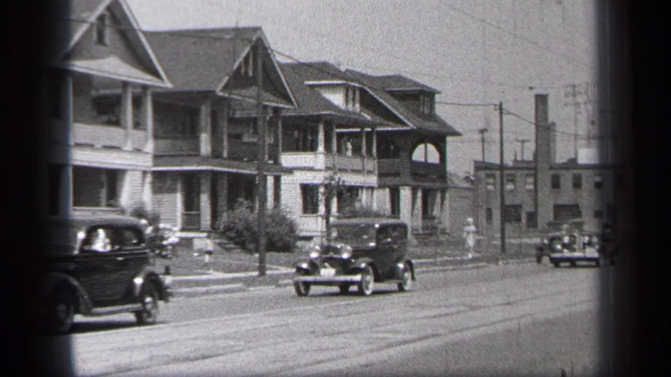 1937 Cleveland frame showing a car crossing the foreground, another car approaching, pedestrians on the sidewalk, and gabled homes behind.