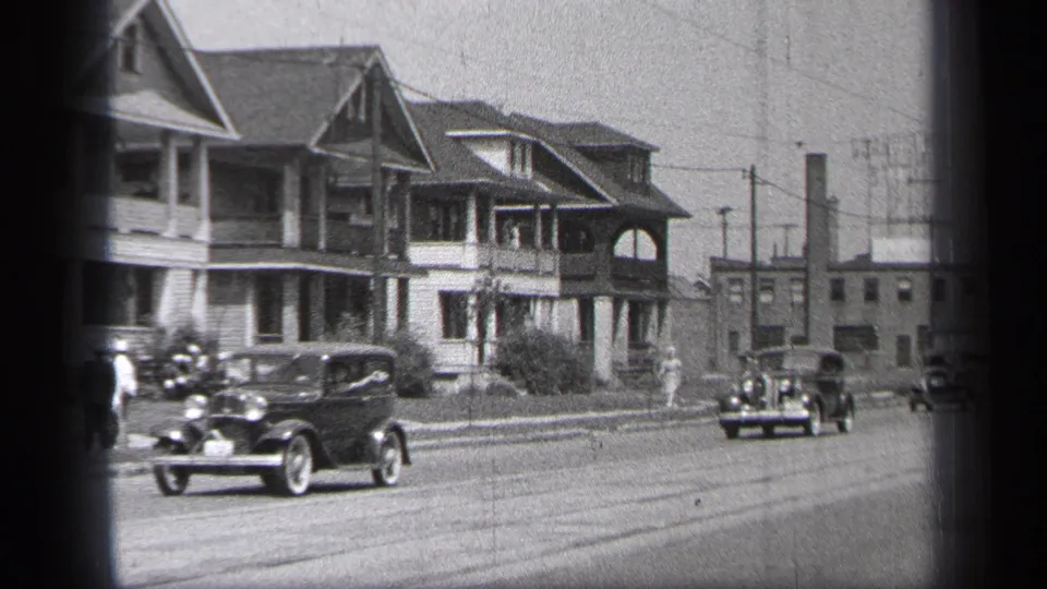 1937 Cleveland home-movie frame with a classic coupe in the foreground, other cars in the distance, porch-front houses, and pedestrians nearby.
