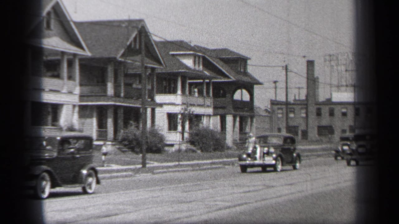 Vintage cars pass two-story porch houses on a city street.