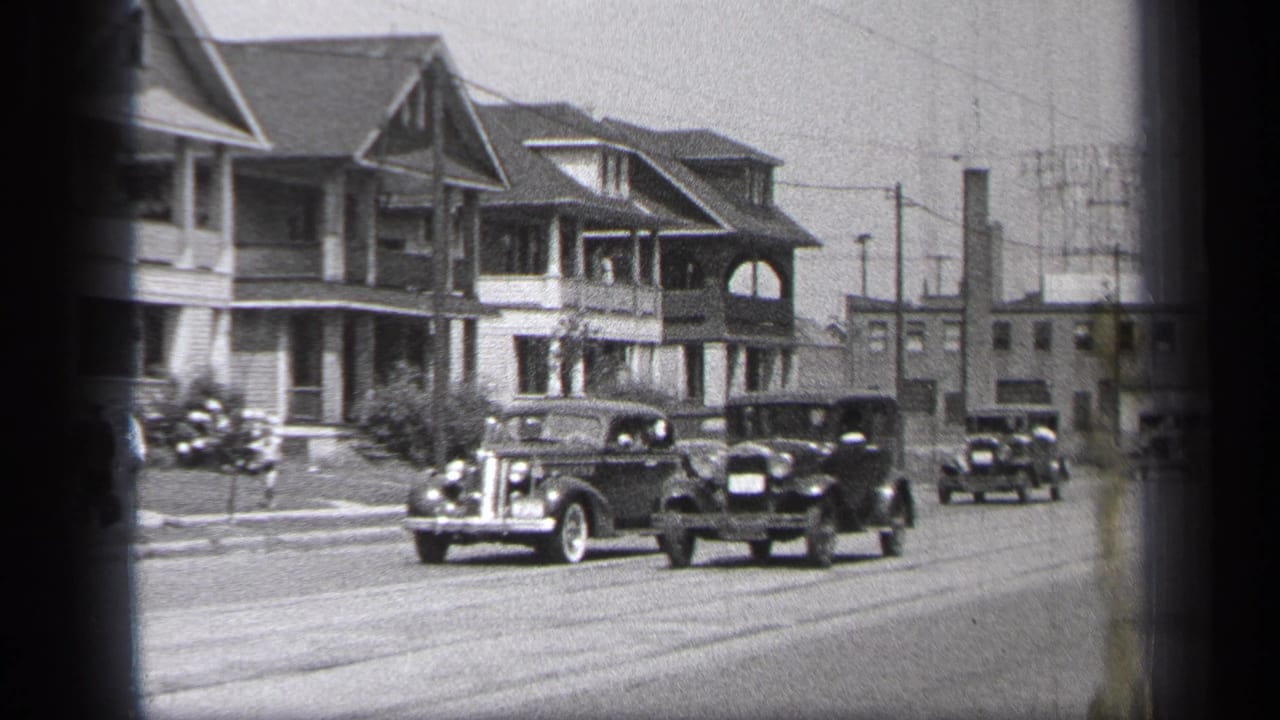 Several old cars move along a residential road beside large houses.