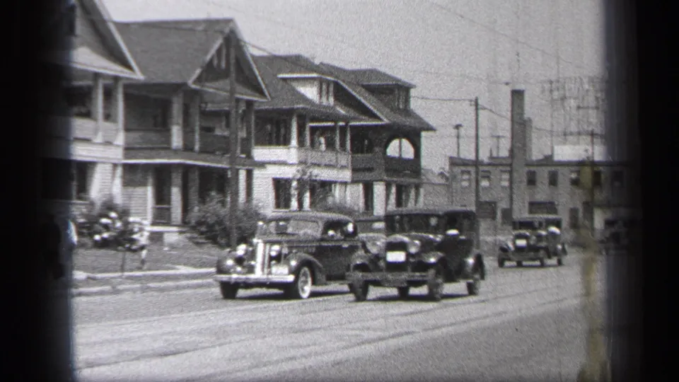 Multiple late-1930s automobiles pass Cleveland houses with porches, with utility poles and low buildings in the background.