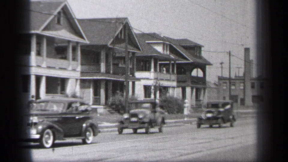 1937 Cleveland curbside view of cars moving past two-story residential houses, with a distant pedestrian near the sidewalk.