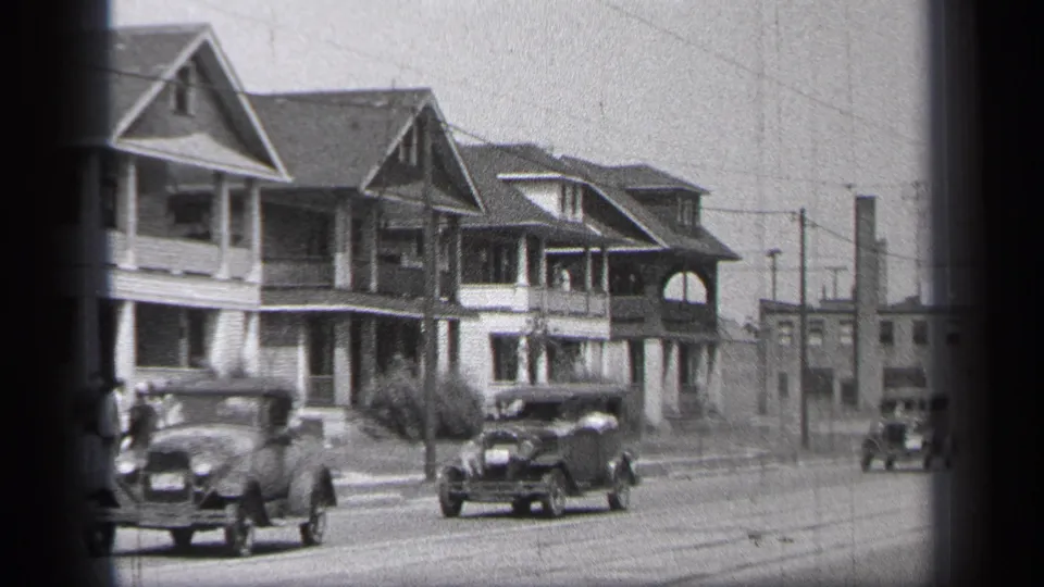 Late-1930s cars travel through a Cleveland residential block with porch houses, wires, and distant low buildings.