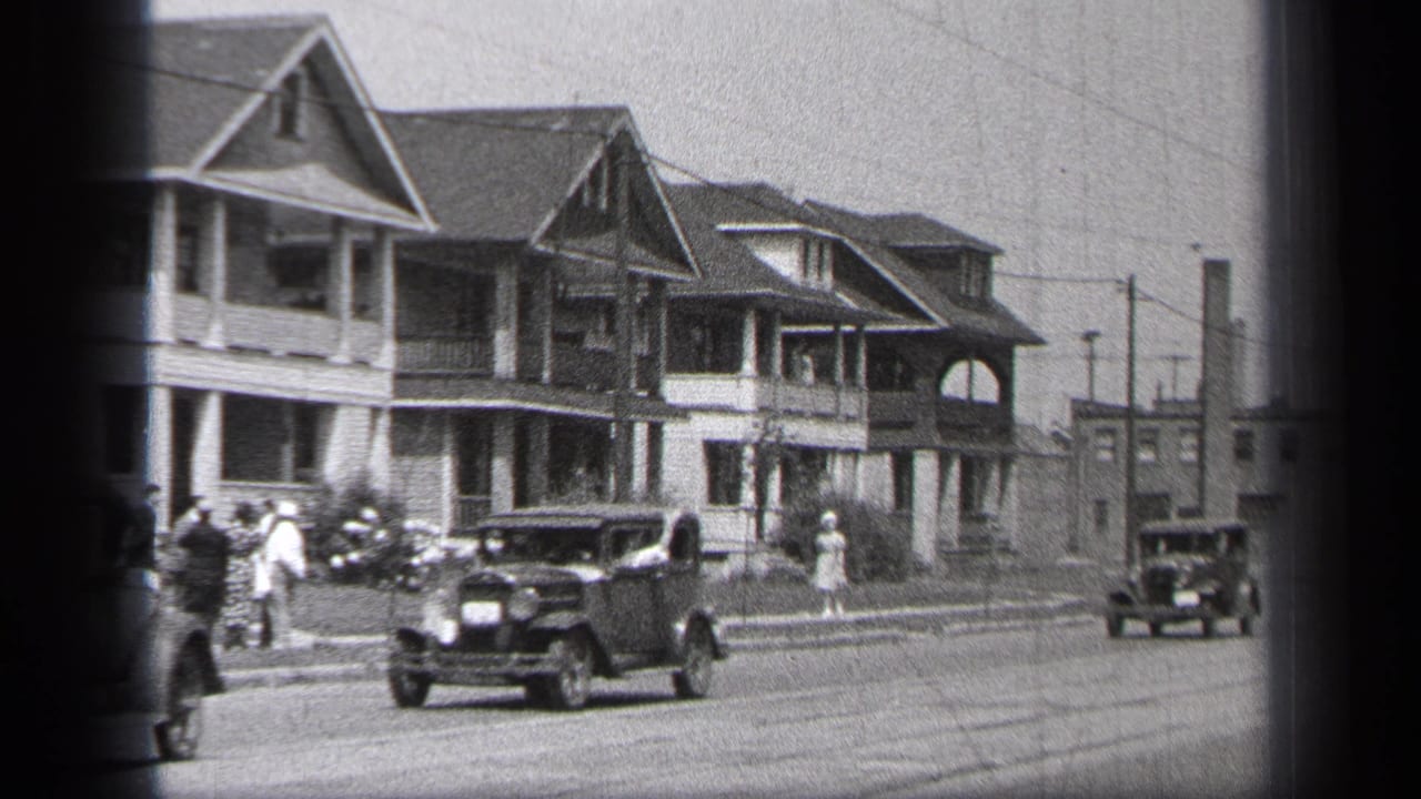 Early automobiles pass large porch-front houses on a residential street with pedestrians nearby.