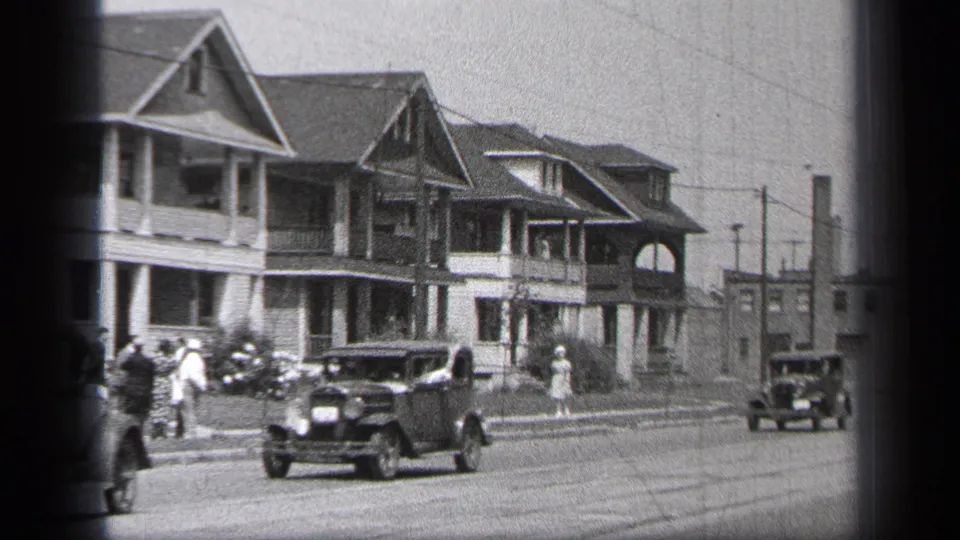 1937 Cleveland home-movie frame showing early cars moving along a residential street lined with large gabled houses, sidewalks, utility poles, and several pedestrians.