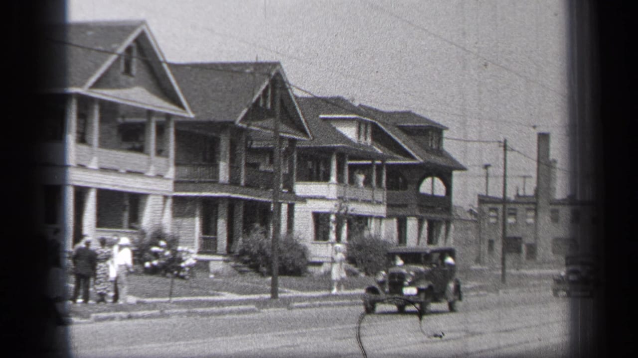 A black automobile drives past two-story houses while people stand near the sidewalk.