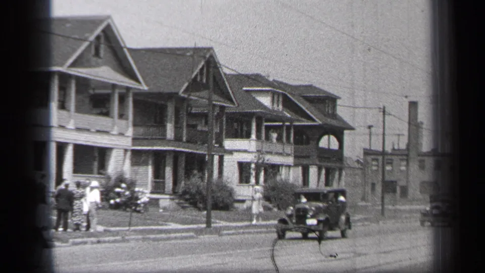 1937 Cleveland residential street view with a dark period automobile, porch-front houses, utility poles, and pedestrians gathered near the curb.