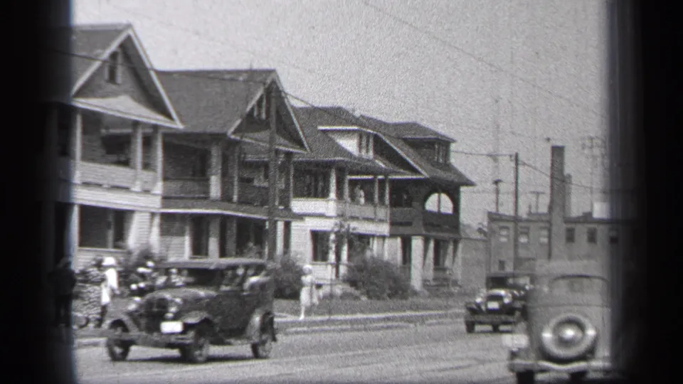 1937 Cleveland street frame showing multiple early automobiles, a row of gabled homes with porches, sidewalk pedestrians, and a partial car entering the foreground.