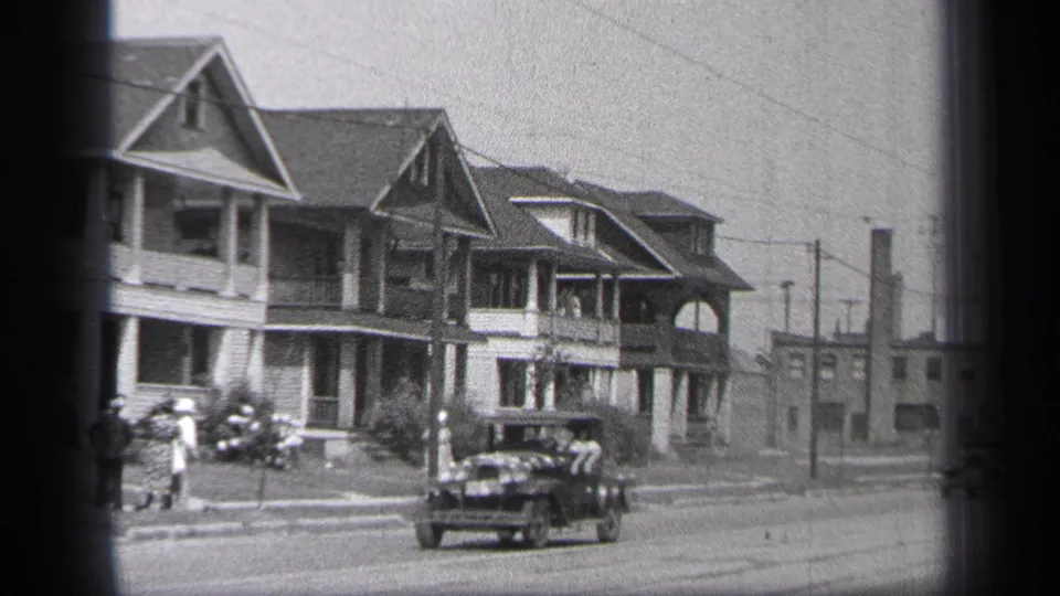 1937 Cleveland home-movie frame with a dark period car centered on the street, large porch-front houses behind it, and pedestrians along the sidewalk.