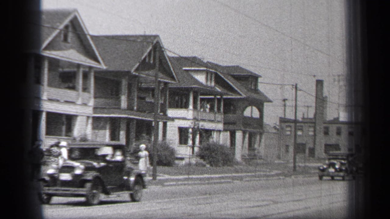 1937 Cleveland home-movie frame showing a dark sedan, sidewalk pedestrians, porch-front houses, and utility poles along a residential street.