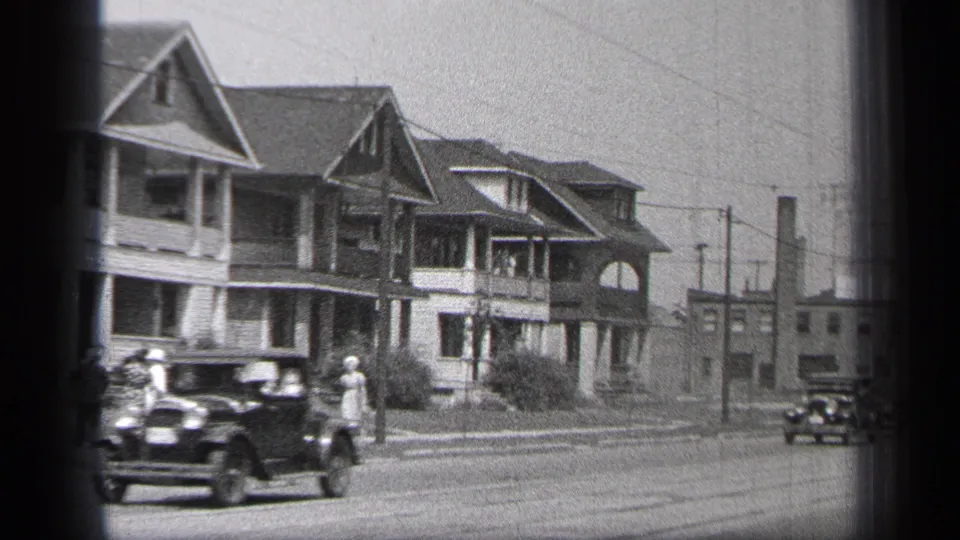 1937 Cleveland home-movie frame showing a dark sedan, sidewalk pedestrians, porch-front houses, and utility poles along a residential street.