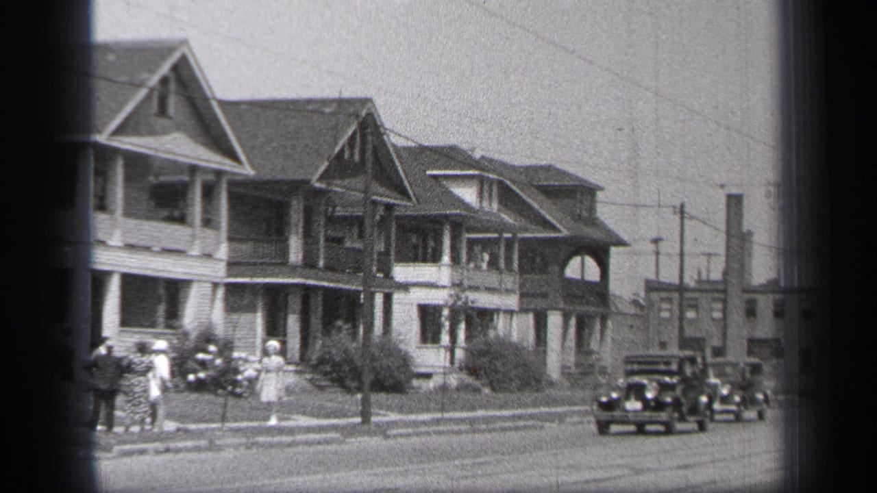Pedestrians stand by the curb as two dark cars travel along a residential street lined with houses.