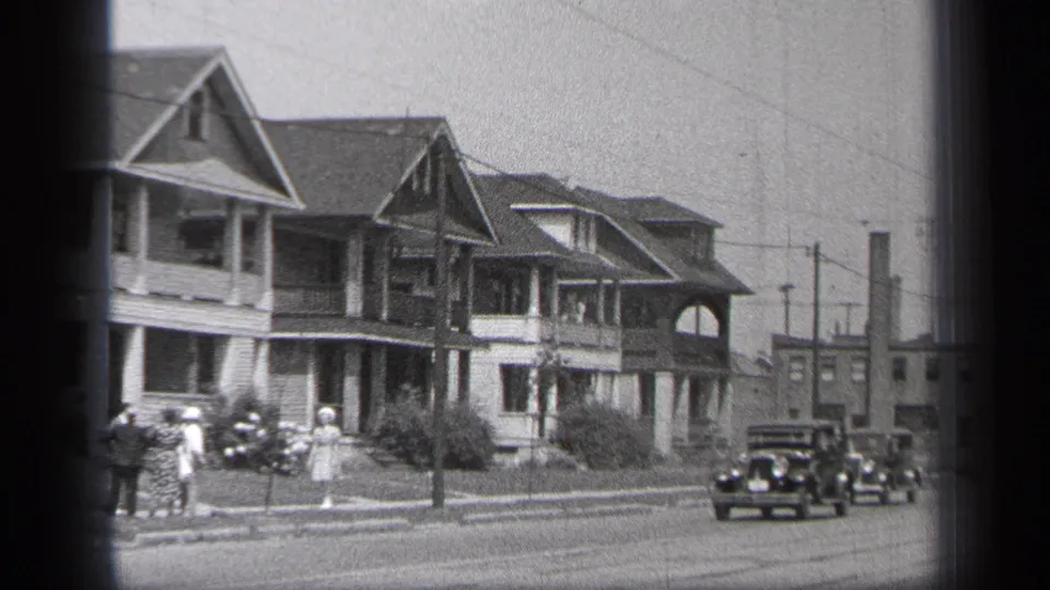 1937 Cleveland street view with curbside pedestrians, passing dark automobiles, porch-front houses, and overhead utility lines.