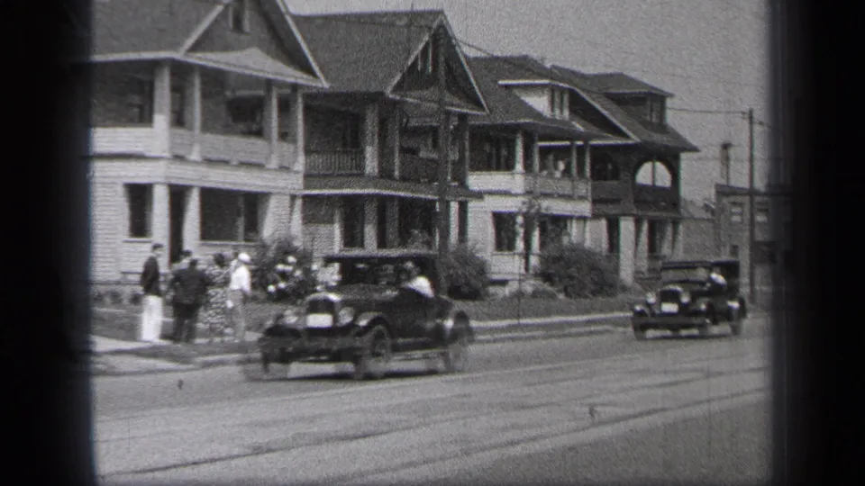 1937 Cleveland frame showing a curbside group, a dark car in motion, residential porches, gabled roofs, and a second car farther down the street.