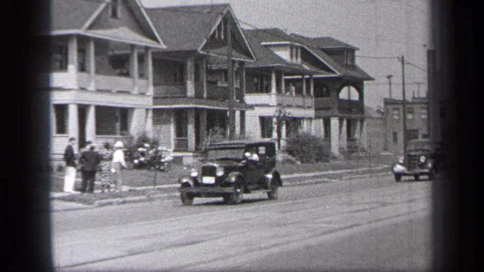 1937 Cleveland home-movie frame with a dark sedan centered on the street, pedestrians at left, residential houses, and a distant car.