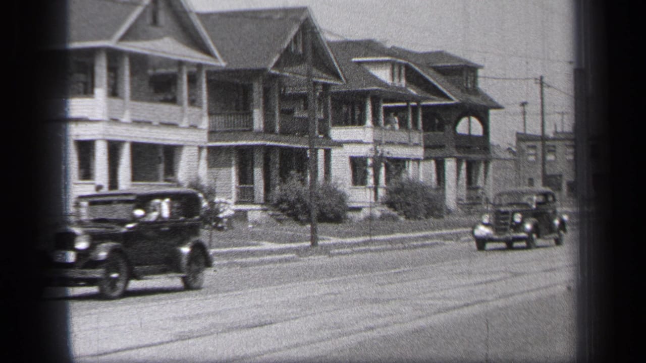 Cars pass a residential street lined with two-story houses and porches.