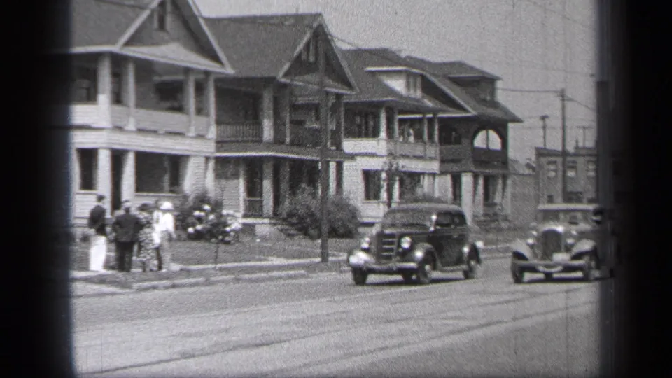 People gather on a Cleveland sidewalk while 1930s cars pass in front of residential houses.
