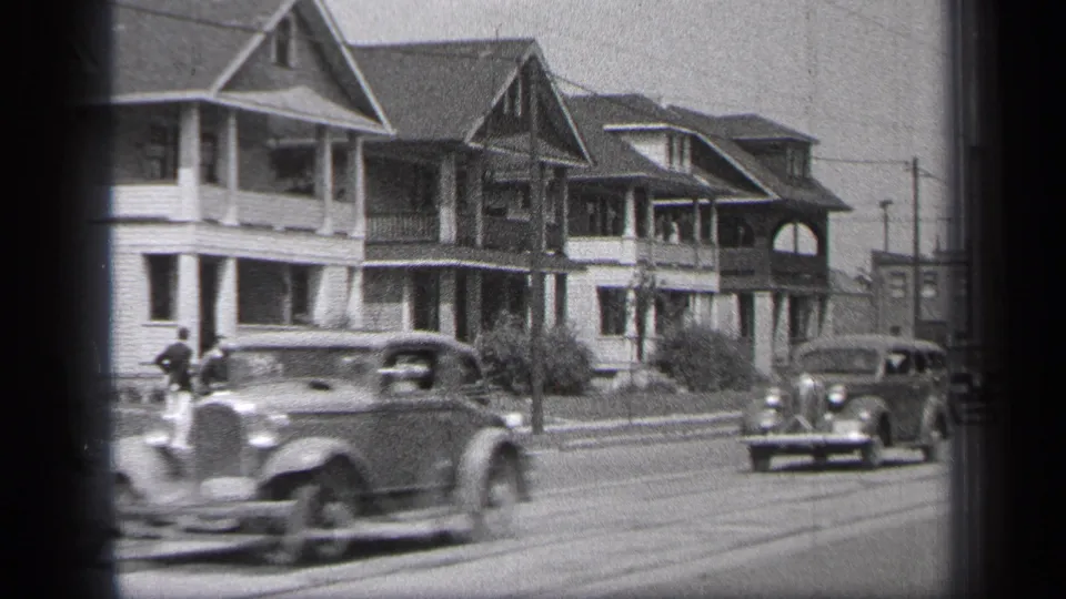 A 1930s automobile blurs past the camera on a Cleveland residential street with gabled homes behind.