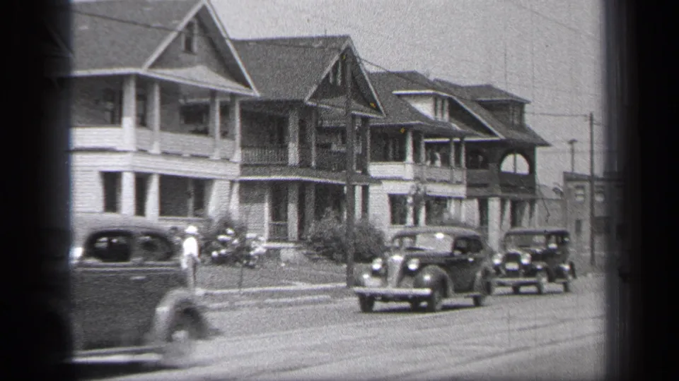 Period cars travel along a 1937 Cleveland residential street bordered by two-story homes and utility poles.