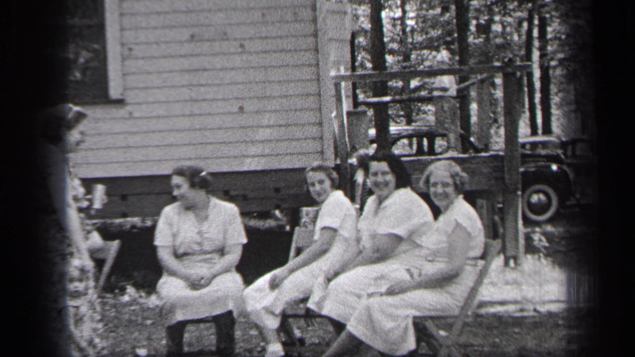 Four women in light dresses sit outdoors beside a wooden porch.
