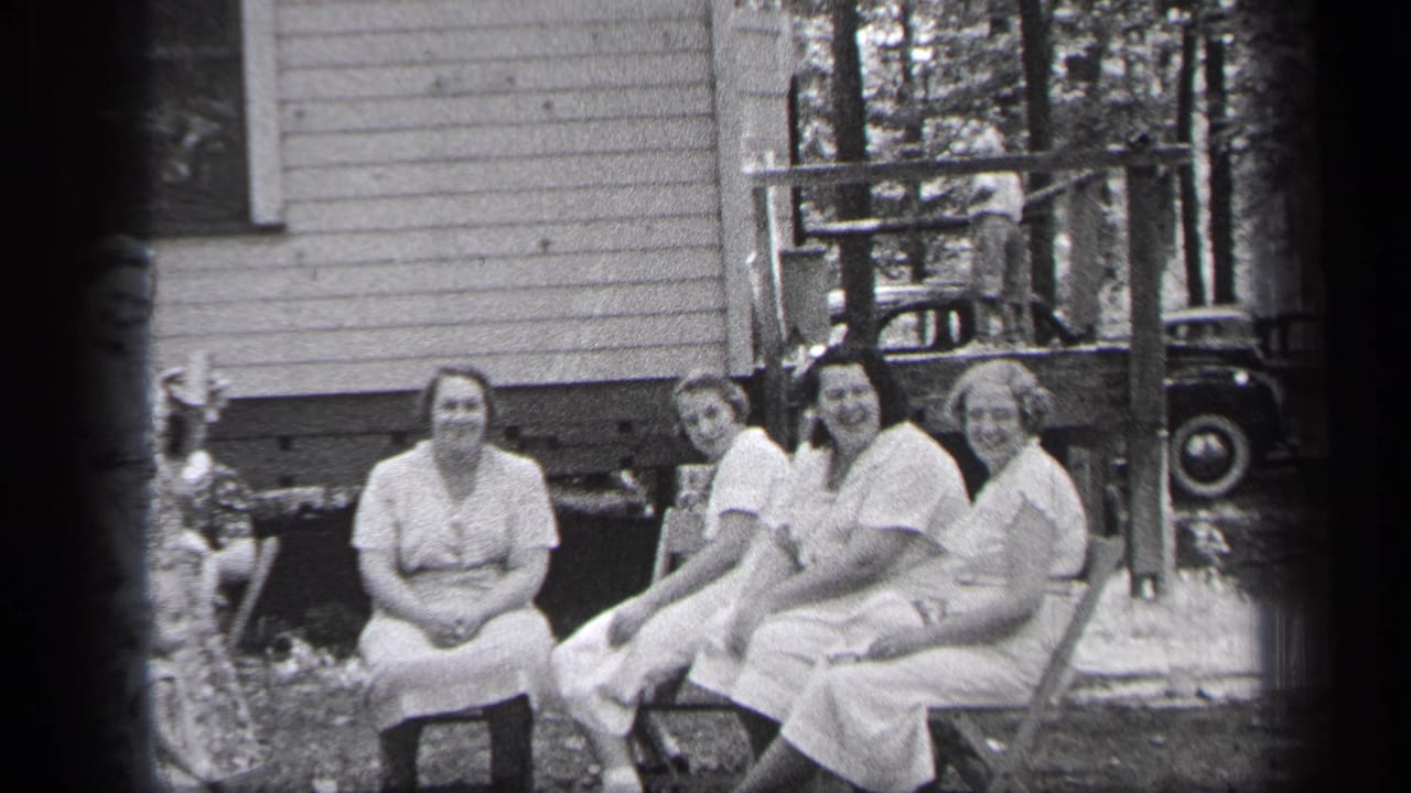 Women sit in a row near a porch, with trees and a car partly visible behind them.