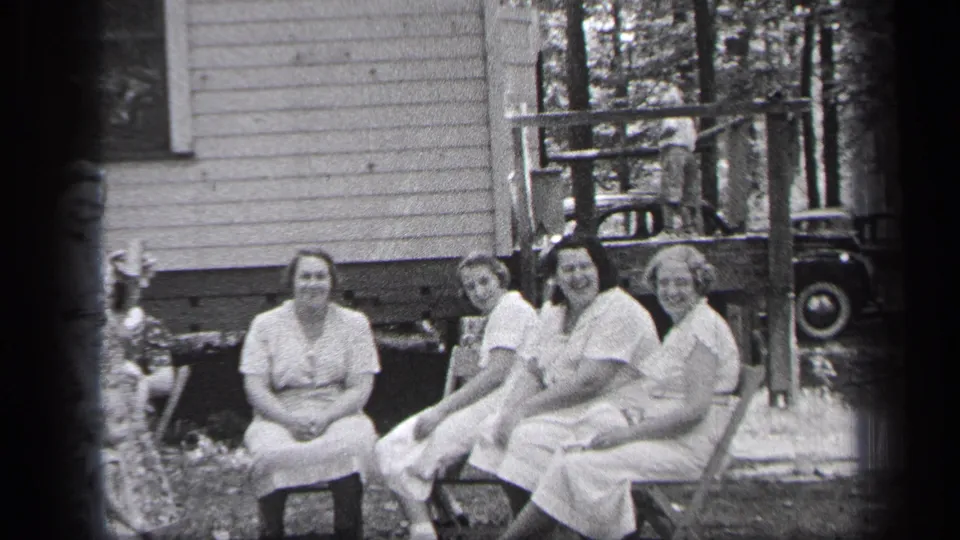Seated women smile near a wooden porch, with trees and a partially visible car in 1937 Cleveland home-movie footage.