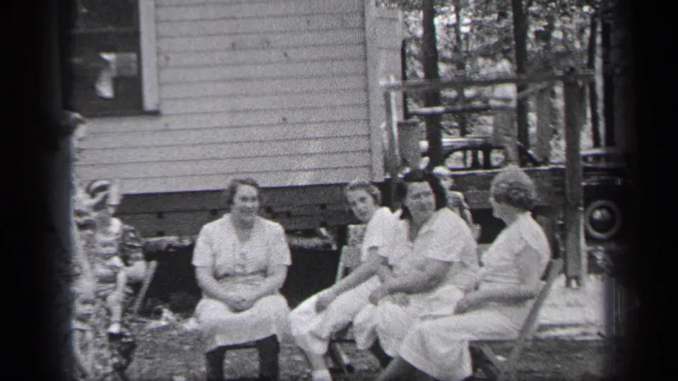 Four women sit outdoors near a porch as a small figure appears at the left edge of the 1937 Cleveland home-movie frame.