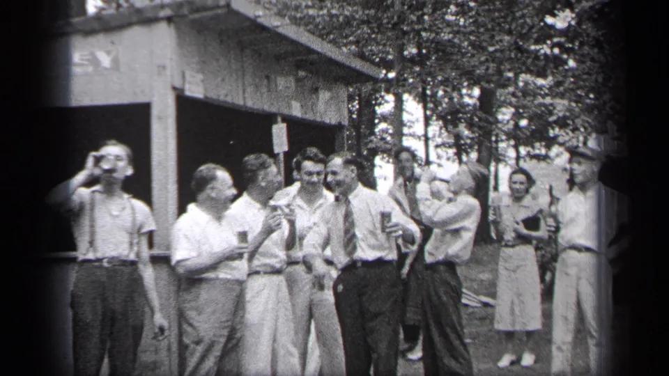 1937 Cleveland frame showing one man lifting a glass as nearby adults converse outside a rustic structure.