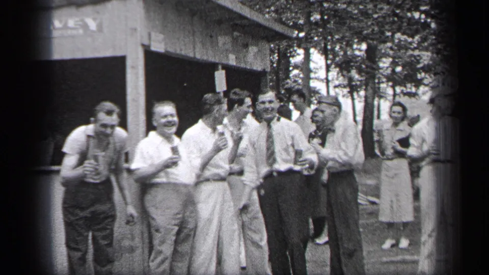 In Cleveland, Ohio, 1937, men in shirts, trousers, and suspenders gather with drinks outside a wooden stand while a woman stands behind them.