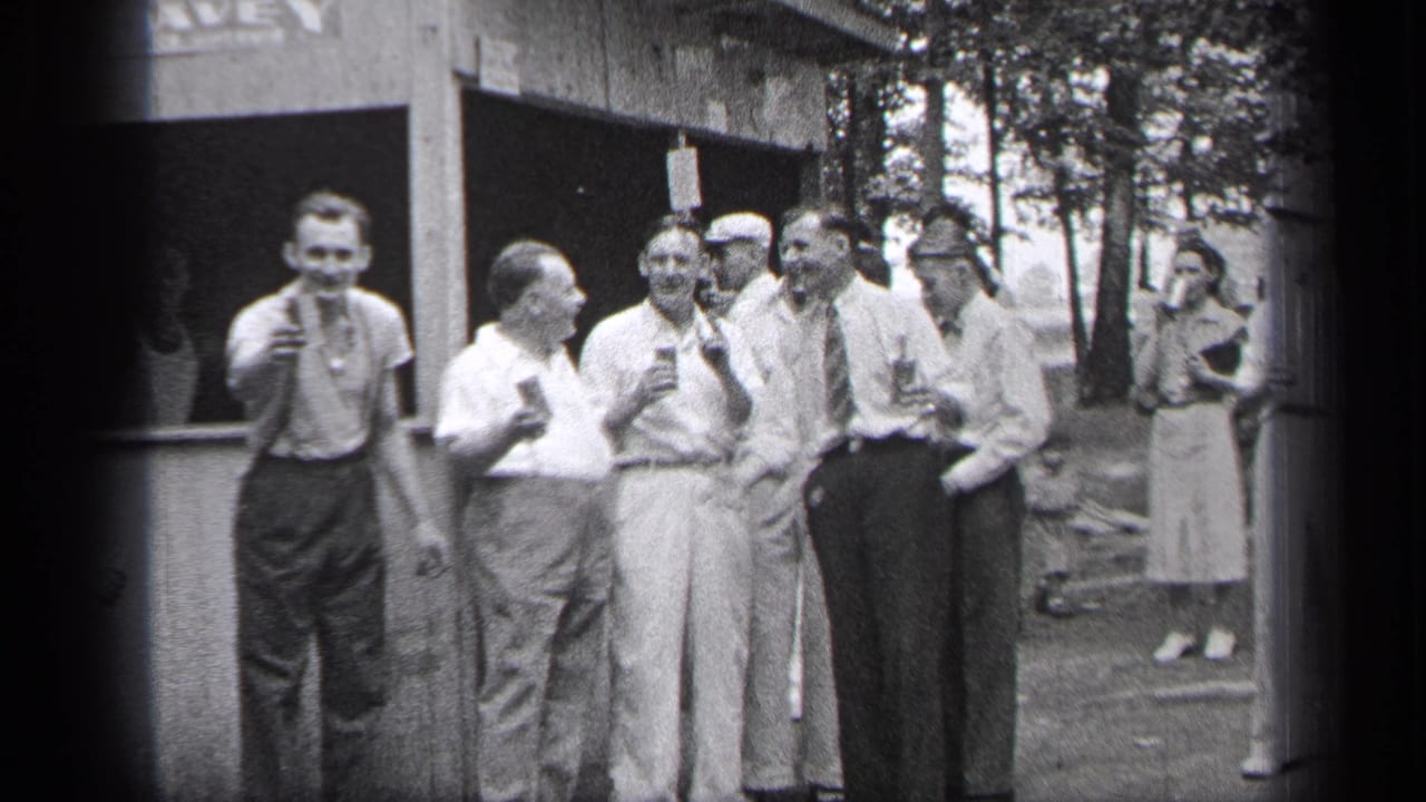 Group of men with drinks stand near a wooden booth as one man faces the camera.