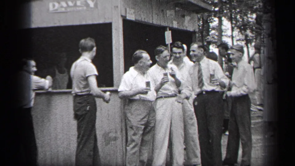 A candid 1937 Cleveland home-movie frame shows men talking with drinks beside a wooden stand, with partial lettering above the opening.