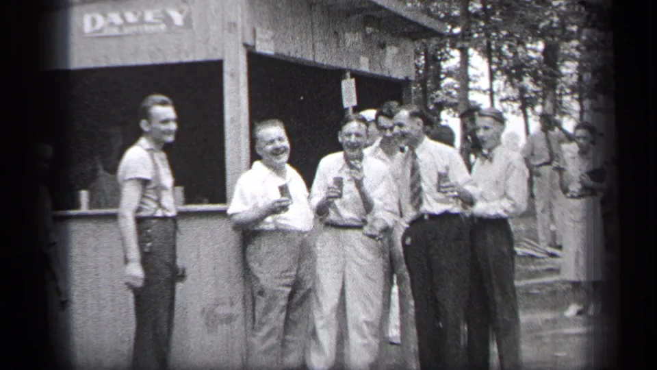 Men in 1930s attire smile and hold drinks near a wooden booth in Cleveland, with trees and a woman in a dress visible behind them.