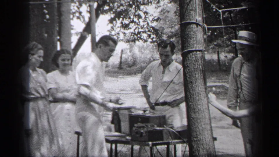 Friends in 1937 Cleveland stand in dappled light around a tabletop record player in a wooded setting.