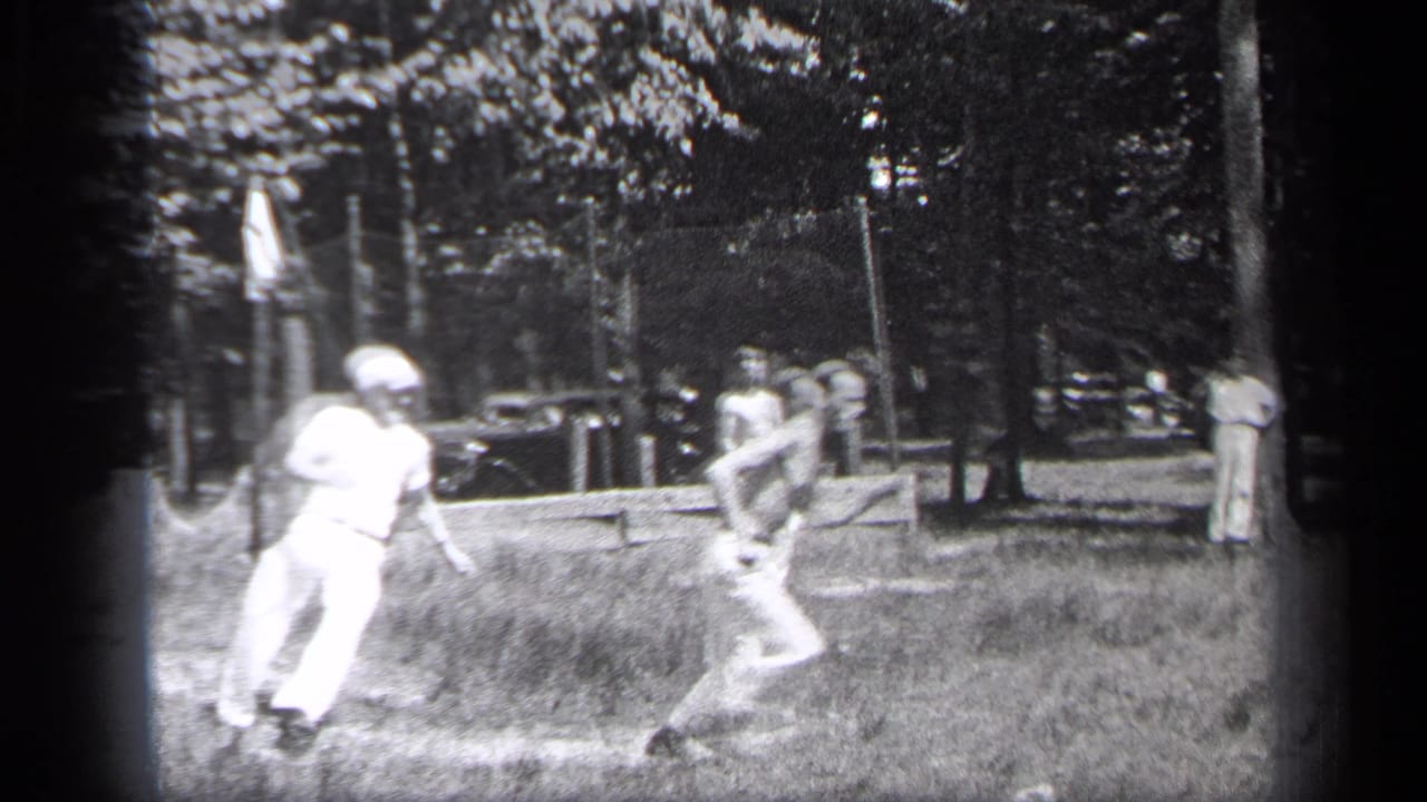 Two men on a grassy field during a casual baseball play, with trees and a parked car behind them.