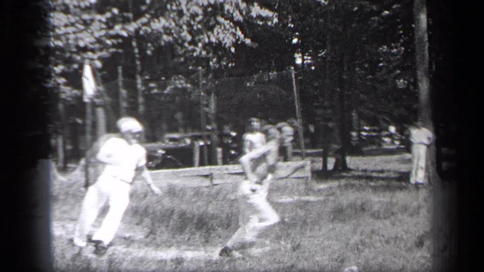 1937 Cleveland home-movie frame of men playing casual baseball in a park, with trees, spectators, and a parked car in the background.
