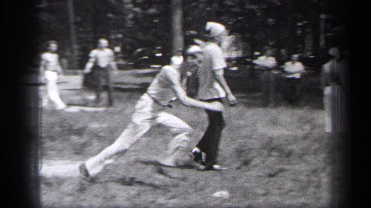Adult men run across a grassy field during a casual ball game as spectators stand nearby.