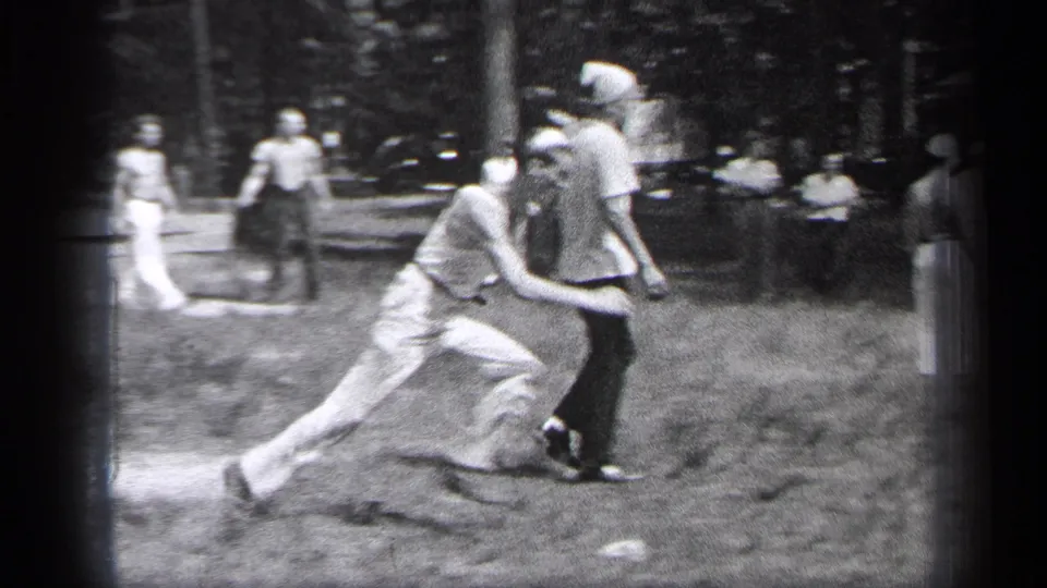 In 1937 Cleveland home-movie footage, adult men in light shirts and trousers chase play on a grassy field with trees and bystanders behind them.
