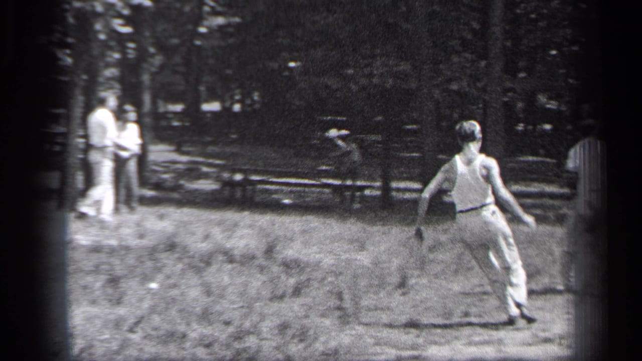 A man runs across the field while other adults stand near trees and benches in the background.