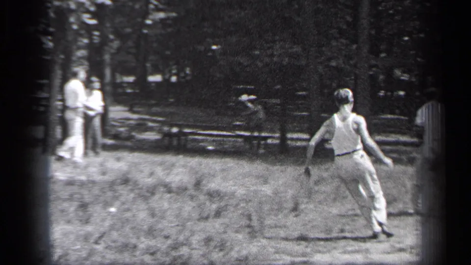 A man in light clothing runs through the grassy playing area in 1937 Cleveland, with adults watching from the shaded park edge.